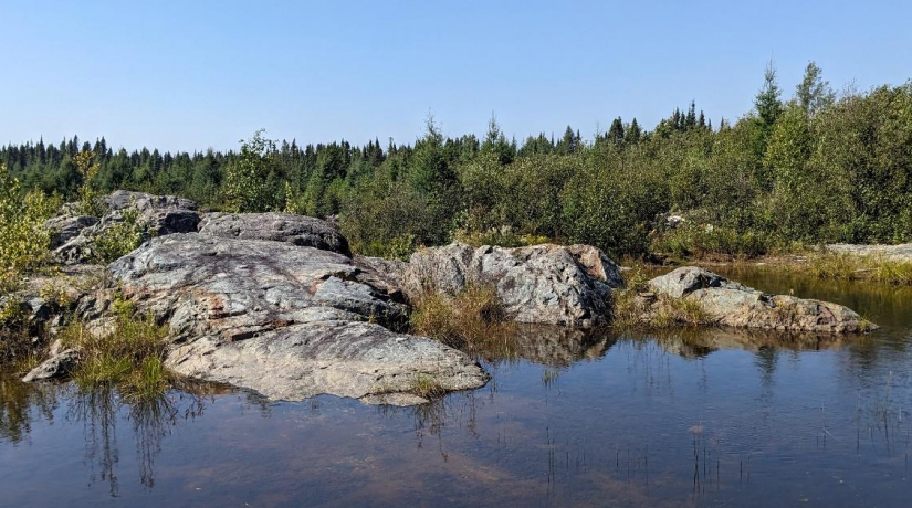 a rock outcrop next to a lake with blue sky and trees in background